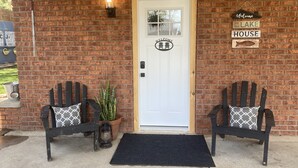 Une entrée de maison en briques avec une porte blanche, deux fauteuils Adirondack dotés de coussins à motifs, une plante en pot, une lanterne et une pancarte de bienvenue.