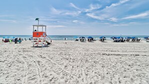 A lifeguard tower on a sandy beach with blue umbrellas and beach chairs in the background.