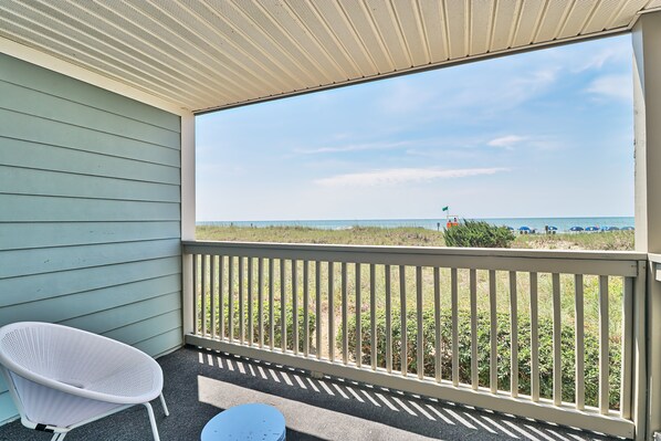 A balcony with a white chair and a small table, overlooking a beach with a fence and distant beachgoers.