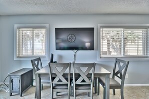 A dining area with a table and four chairs, a television mounted on the wall, and two windows with blinds.
