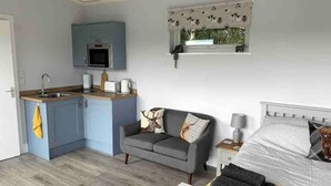 Un espace de vie compact comprenant un coin cuisine, un canapé gris agrémenté de coussins décoratifs, un lit blanc avec une couverture à motifs et un petit tapis blanc.
