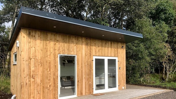 Une cabane moderne en bois, dotée d’une terrasse, de grandes portes-fenêtres coulissantes et d’un toit noir.
