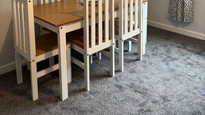 Un comedor con una mesa de madera y seis sillas blancas, un televisor de pantalla plana montado en la pared y una cortina estampada en la ventana.