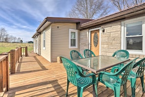 A wooden deck with a glass table and green chairs, leading to a house with a stone exterior and a wooden door.
