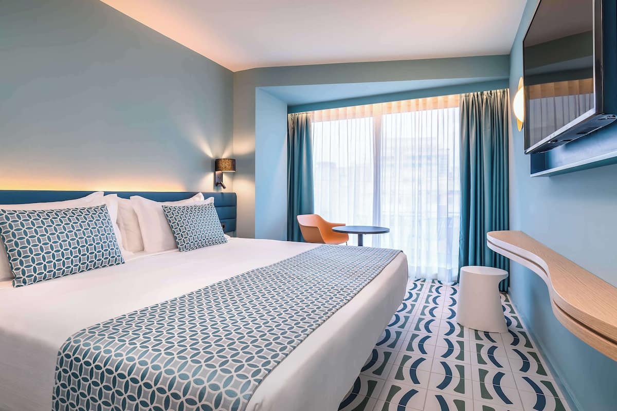 A hotel room with a large bed, a small table, a chair, and a TV mounted on the wall.