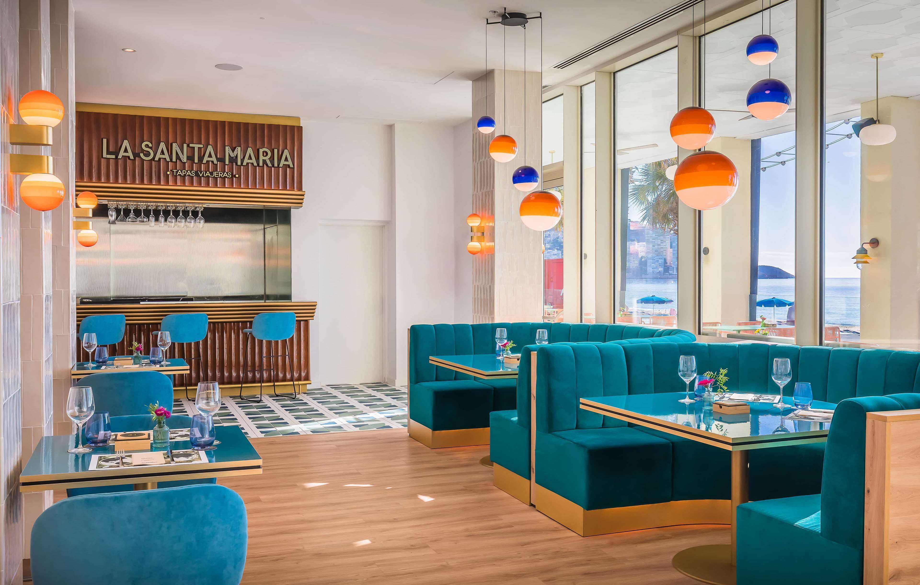 Un restaurante con asientos y mesas color turquesa, una zona de bar y ventanas amplias que ofrecen vistas.