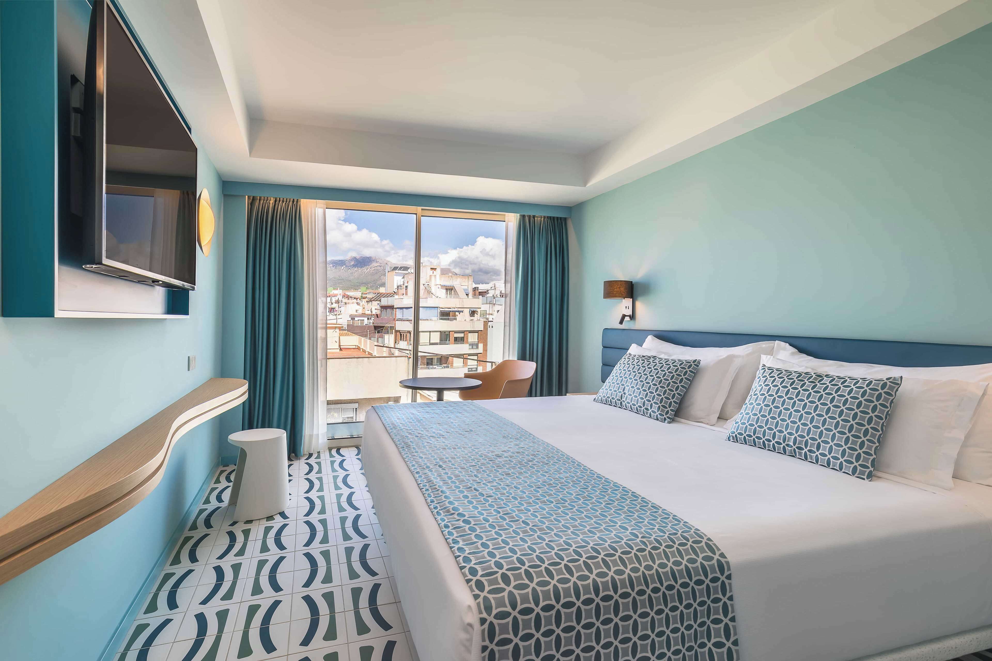 A hotel room with a large bed, a TV mounted on the wall, a small table, and a view of the city through the window.