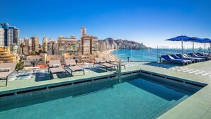 Piscina en la azotea con sillones de descanso y sombrillas, con vistas a una playa y al horizonte urbano.