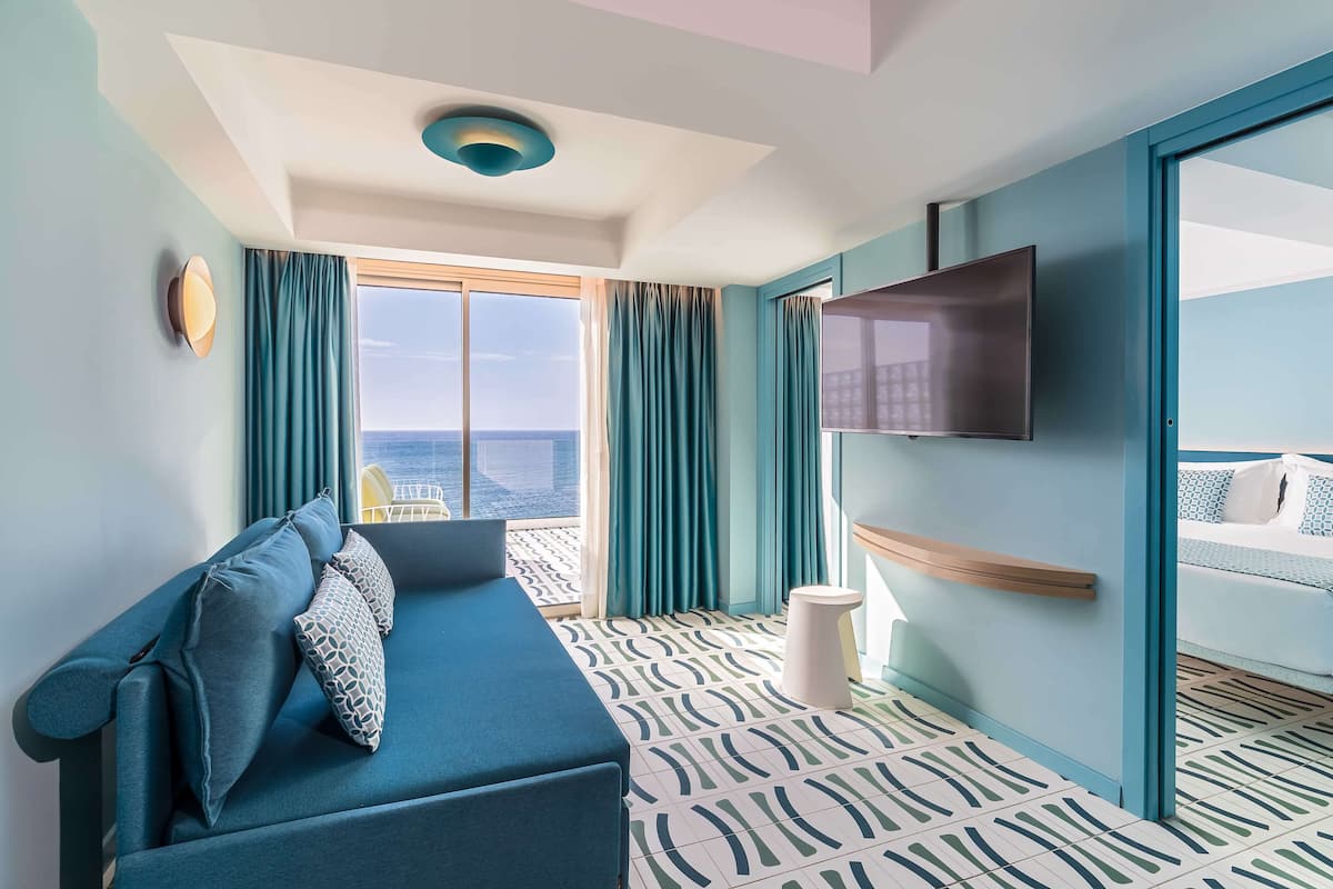 A modern hotel room with a large window, a sofa, a flat-screen TV, and a bed with a blue and white patterned bedspread.