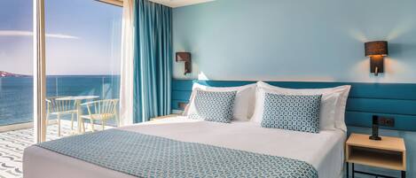 Habitación de hotel con una cama grande, dos lámparas fijadas a la pared, una mesita de noche y vista al mar a través de una puerta corrediza de vidrio.