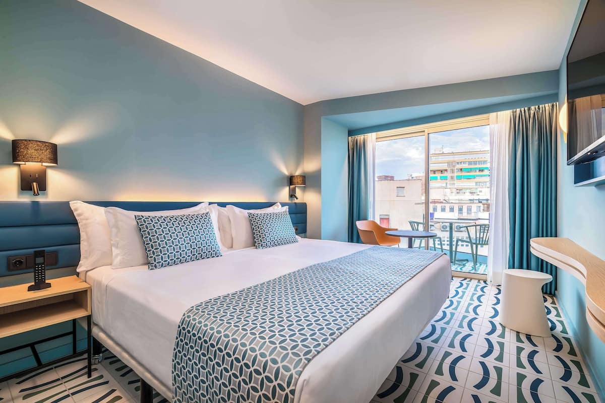 A hotel room with a large bed, a bedside table with a lamp, a small table and chair by the window, and a view of the city.