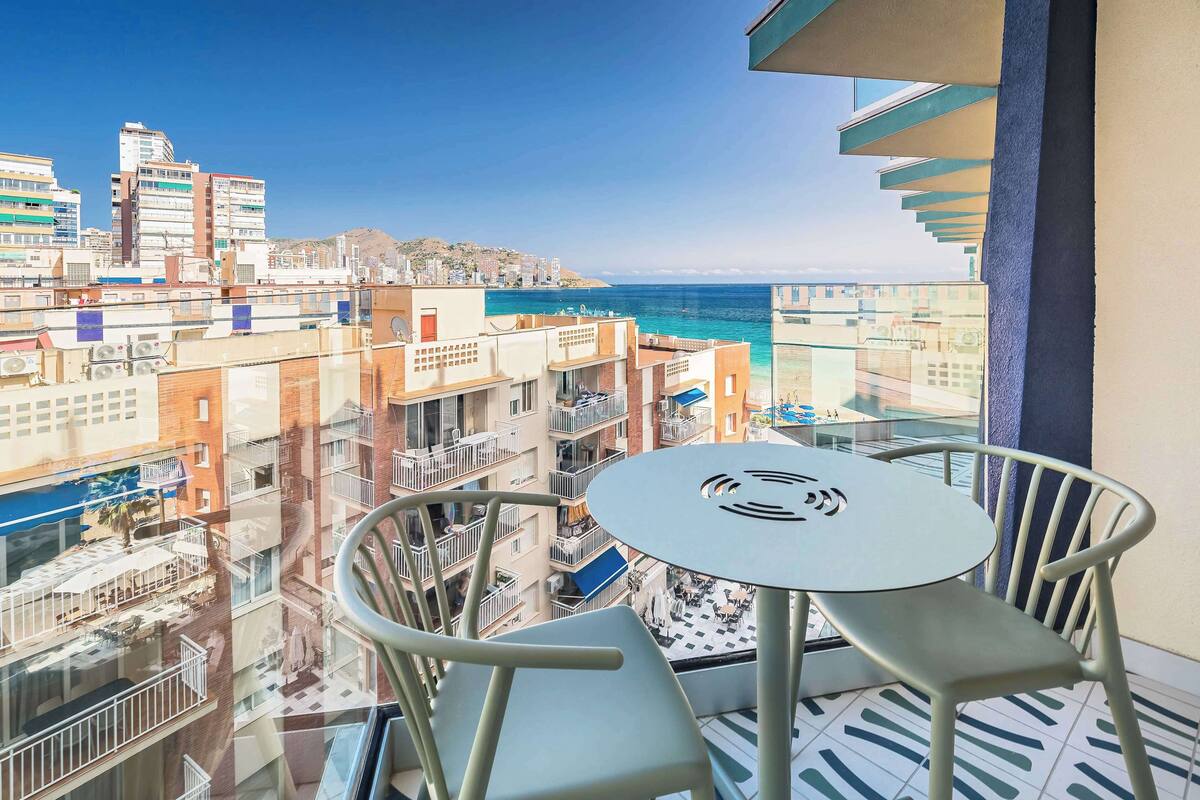 A balcony with a table and chairs overlooking a cityscape and the sea.