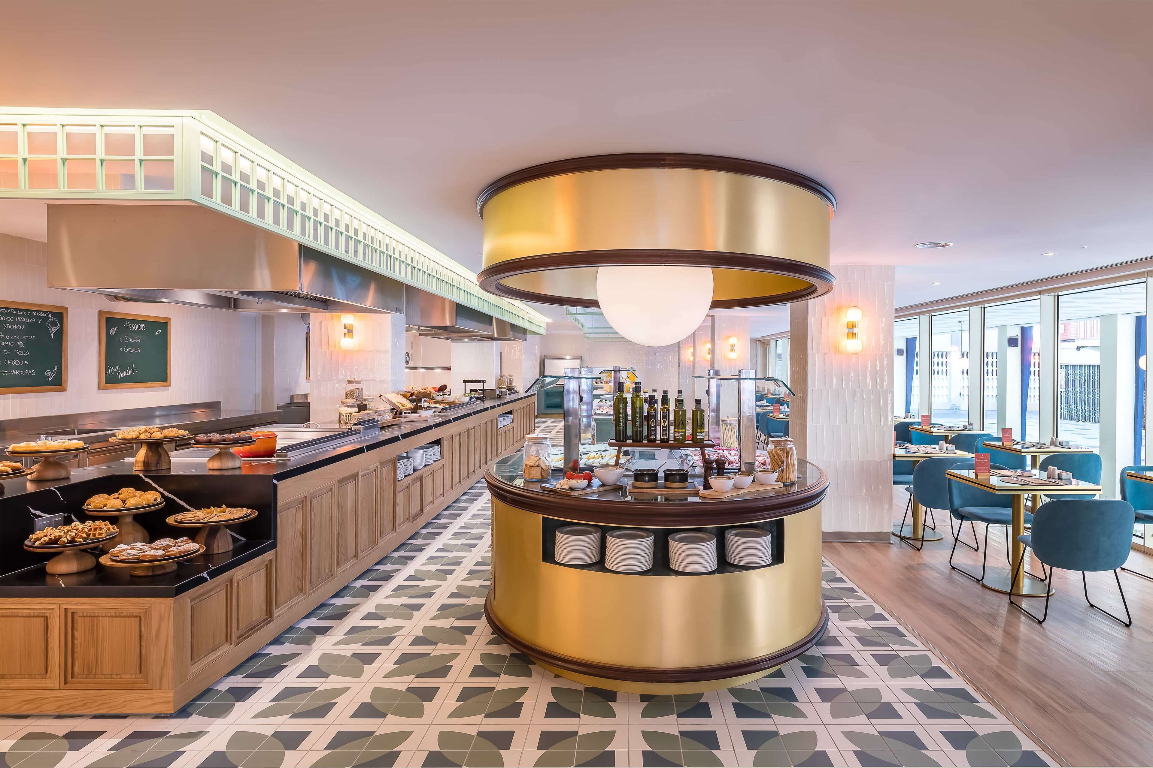 A modern hotel lobby with a central bar area, buffet with various food items, and seating arrangements.