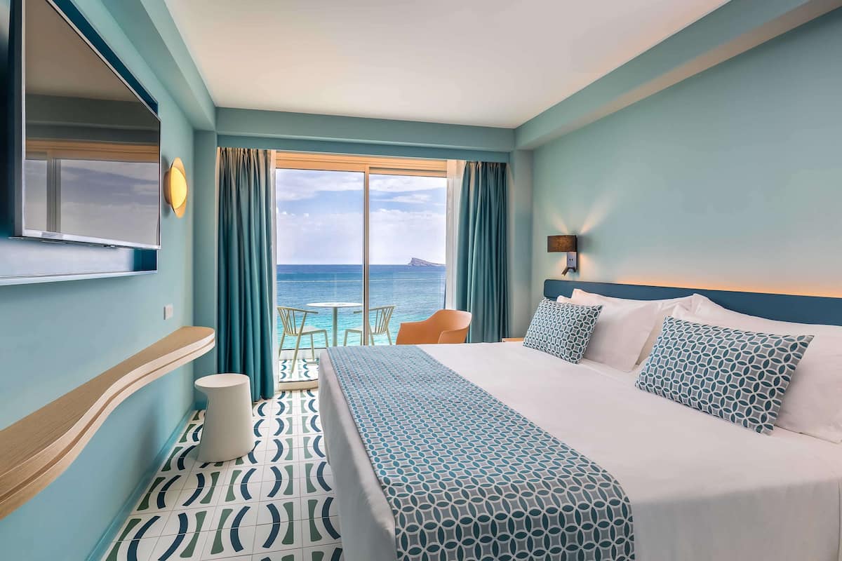 A hotel room with a large bed, a TV, a view of the sea, and a small table with chairs.