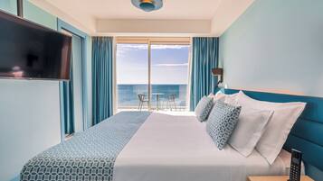 A hotel room with a large bed, a TV, a bedside table with a phone, and a view of the ocean through a large window.