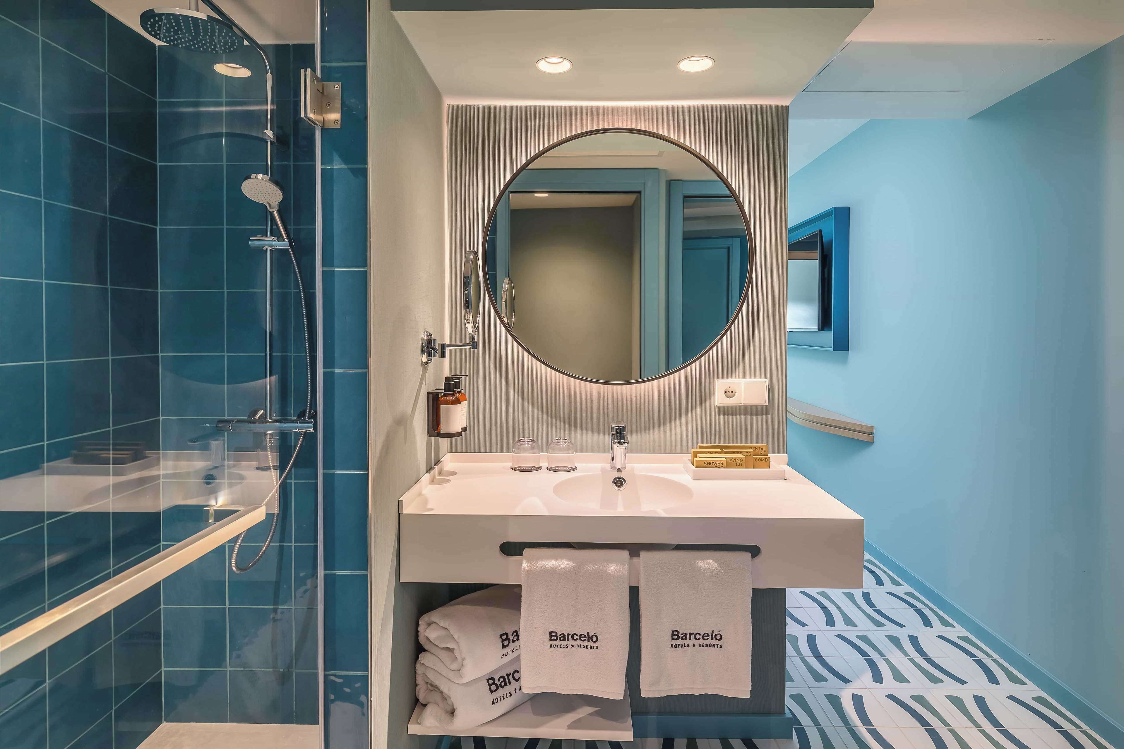 A modern bathroom with a shower, a sink, and a mirror. There are towels with the hotel name on them.
