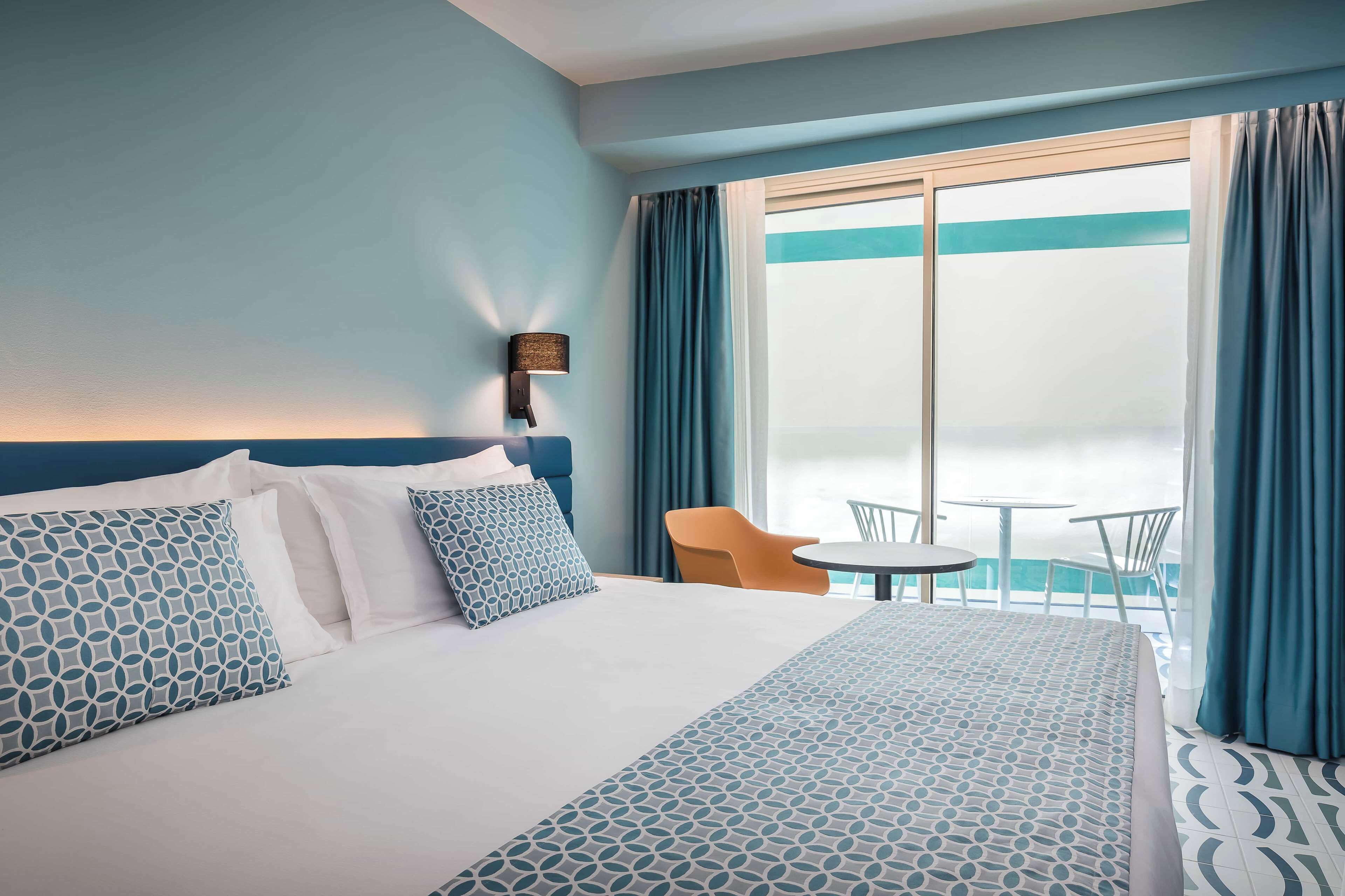 A hotel room with a bed, a nightstand, a lamp, a window with curtains, and a balcony with a table and chairs.