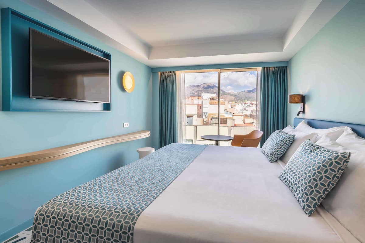 A hotel room with a large bed, a TV mounted on the wall, a balcony with a view of buildings, and a small table with a chair.