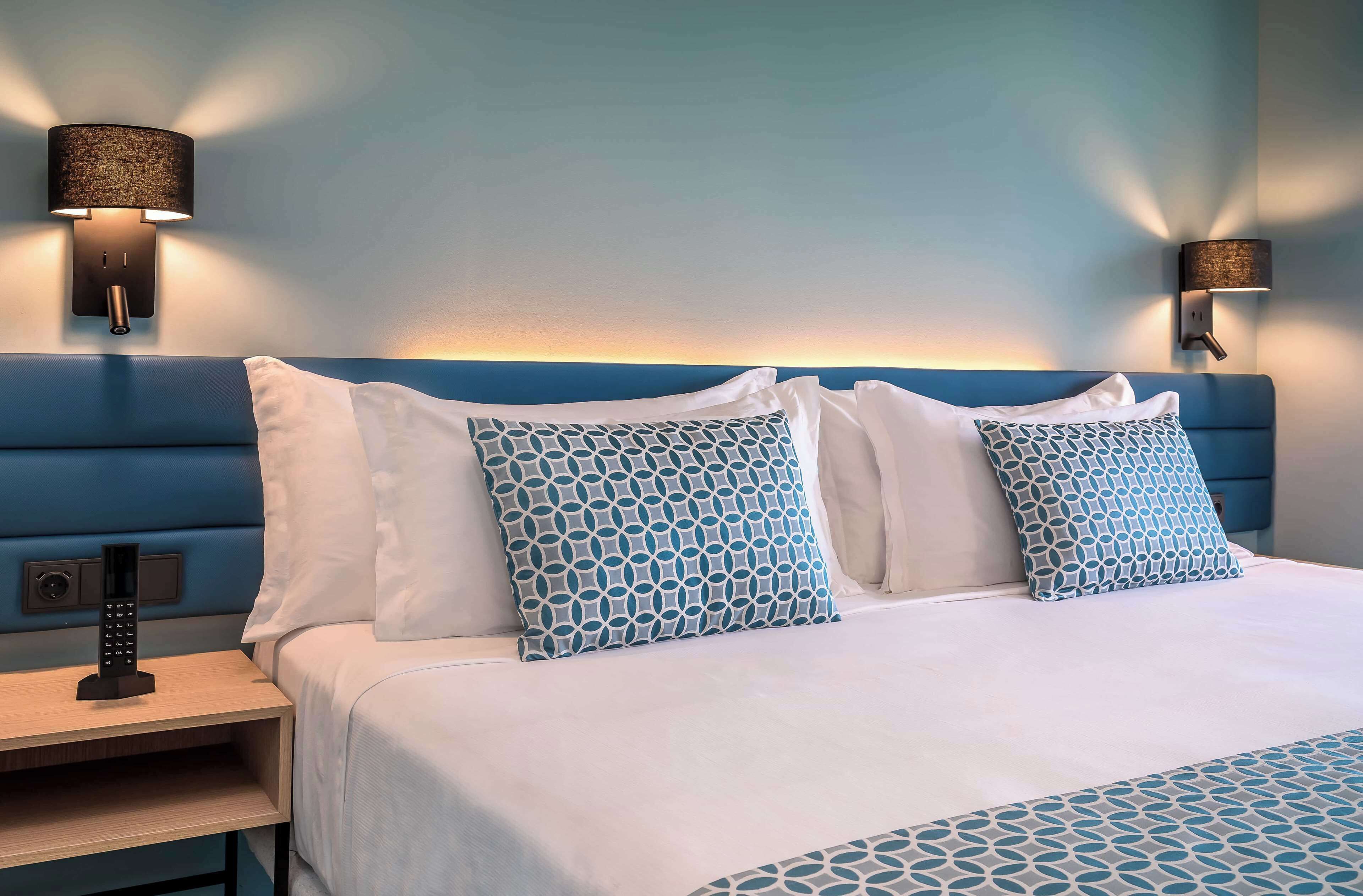 Una cama bien hecha con almohadones estampados, una mesita de noche con control remoto y lámparas fijadas a la pared.