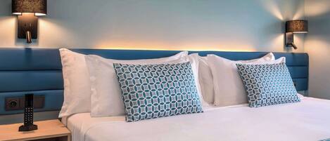 Una cama bien hecha con almohadones estampados, una mesita de noche con control remoto y lámparas fijadas a la pared.
