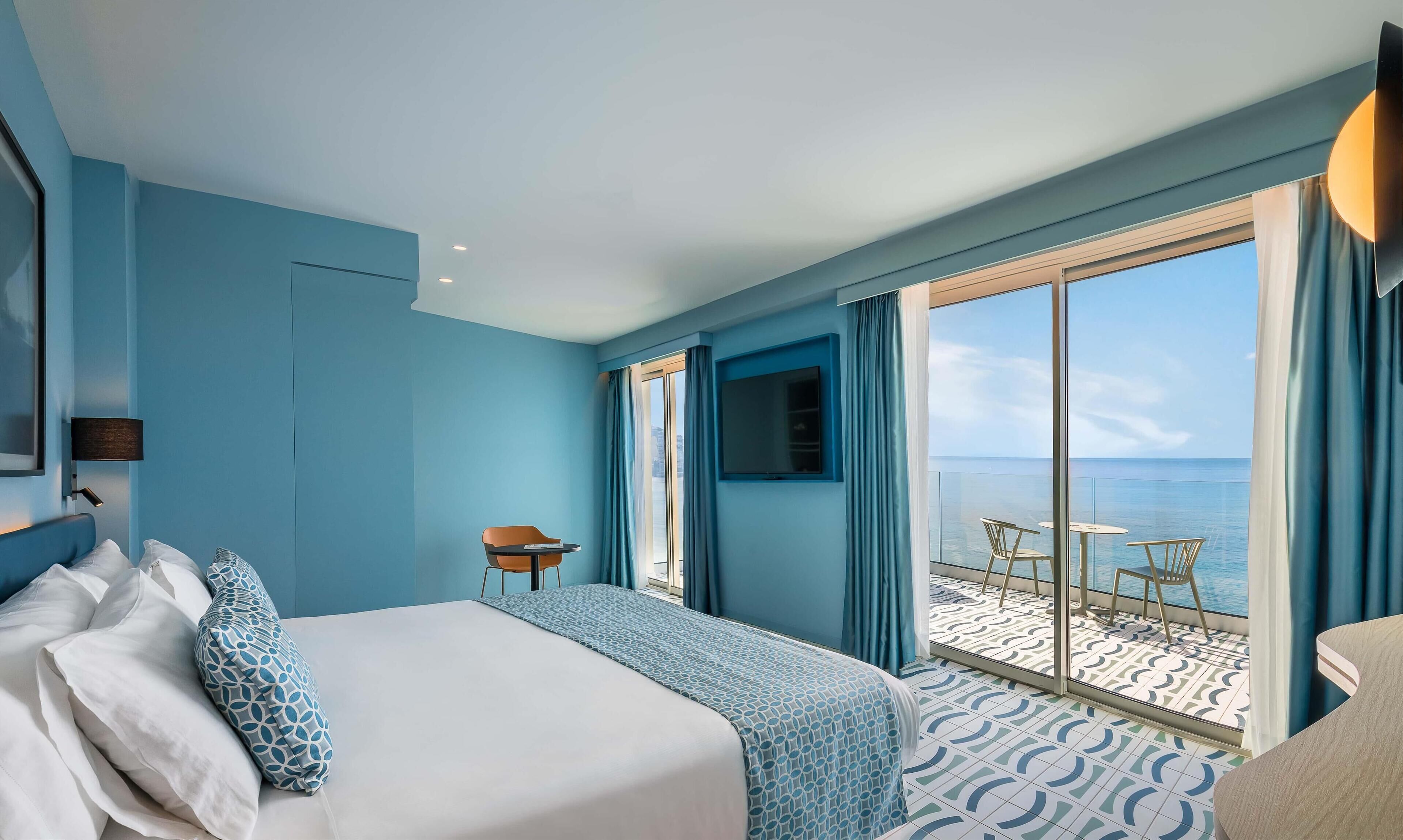 A hotel room with a bed, a chair, a small table, and a balcony with a view of the sea.