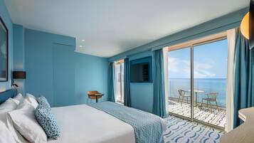 A hotel room with a bed, a chair, a small table, and a balcony with a view of the sea.