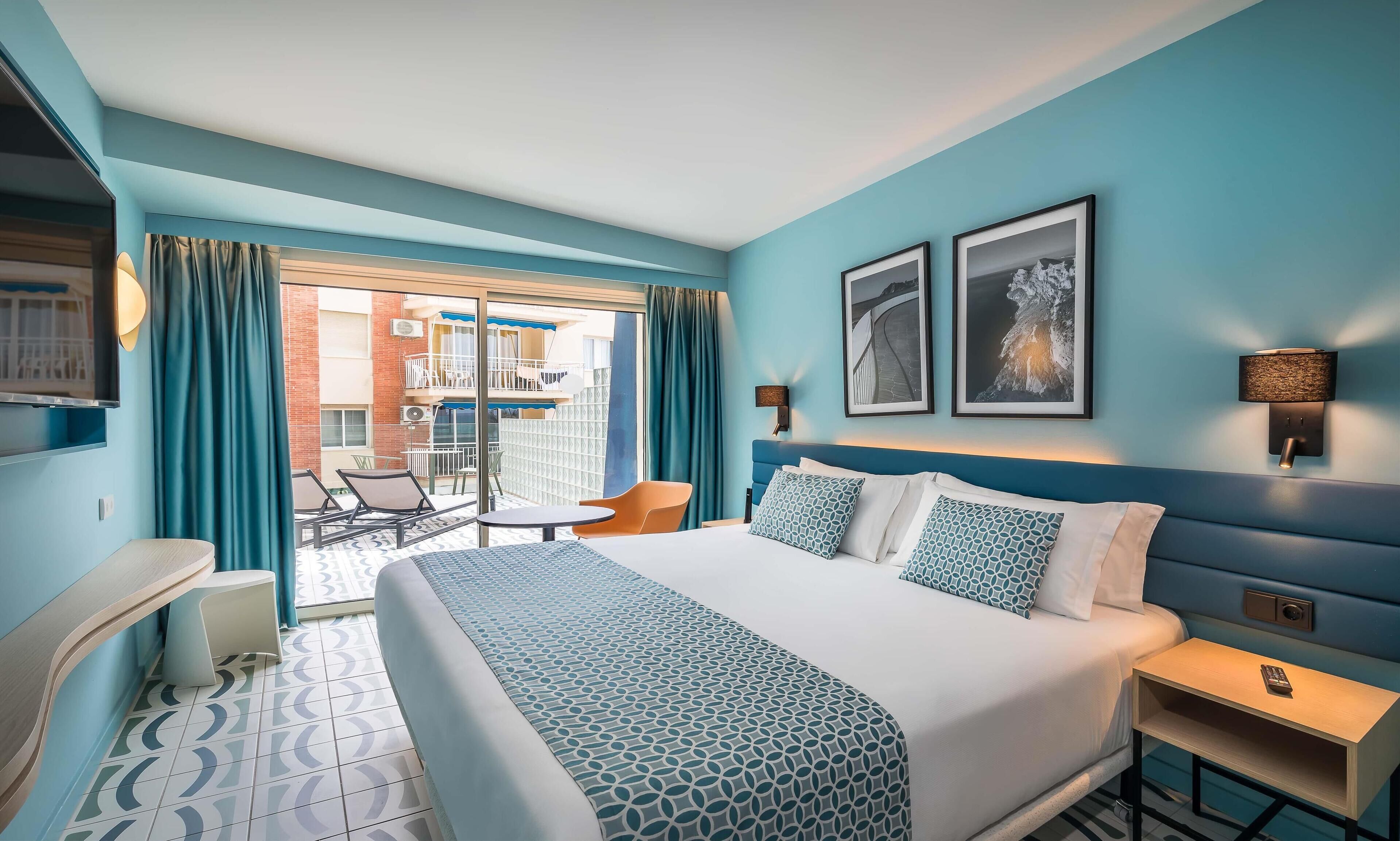 A hotel room with a bed, a desk, a chair, and a balcony with a view of the building.