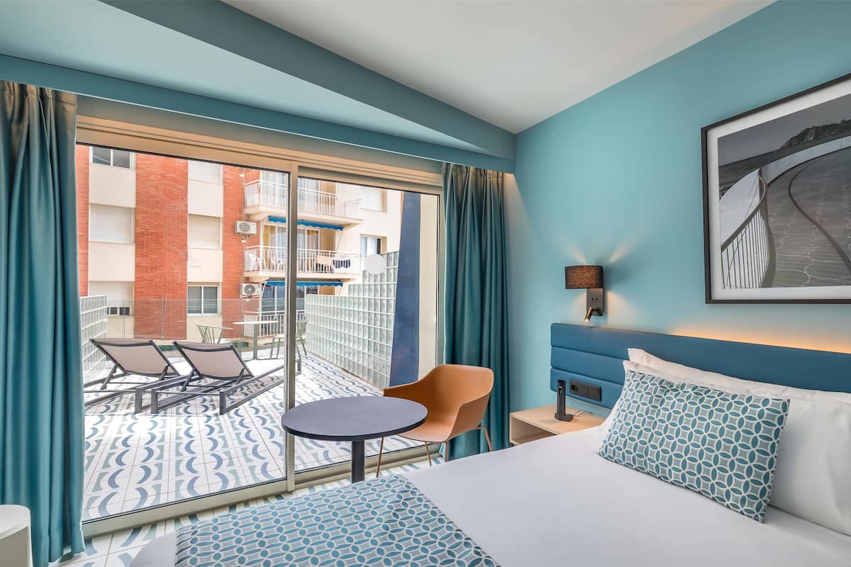A hotel room with a bed, a small table, a chair, a sofa, a balcony with a view of the building, and a framed picture on the wall.