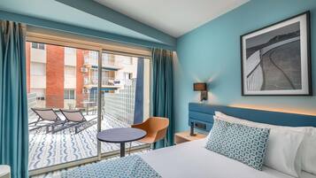 A hotel room with a bed, a small table, a chair, a sofa, a balcony with a view of the building, and a framed picture on the wall.