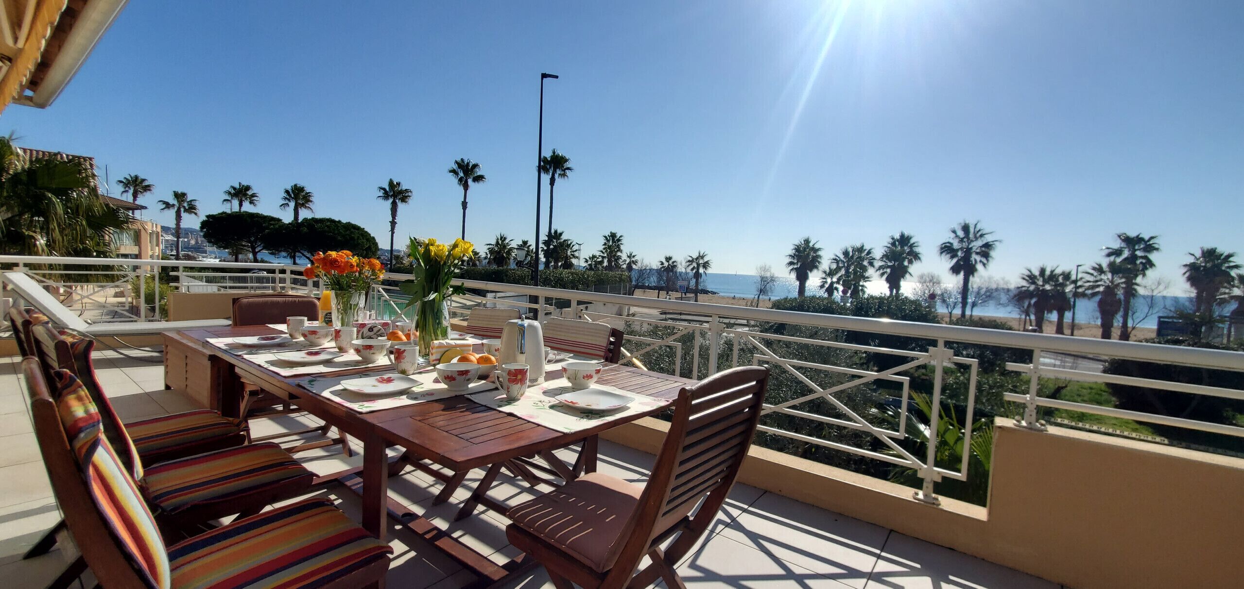 Una terrazza con un tavolo apparecchiato per mangiare, con vista su una zona costiera con palme e cielo sereno.