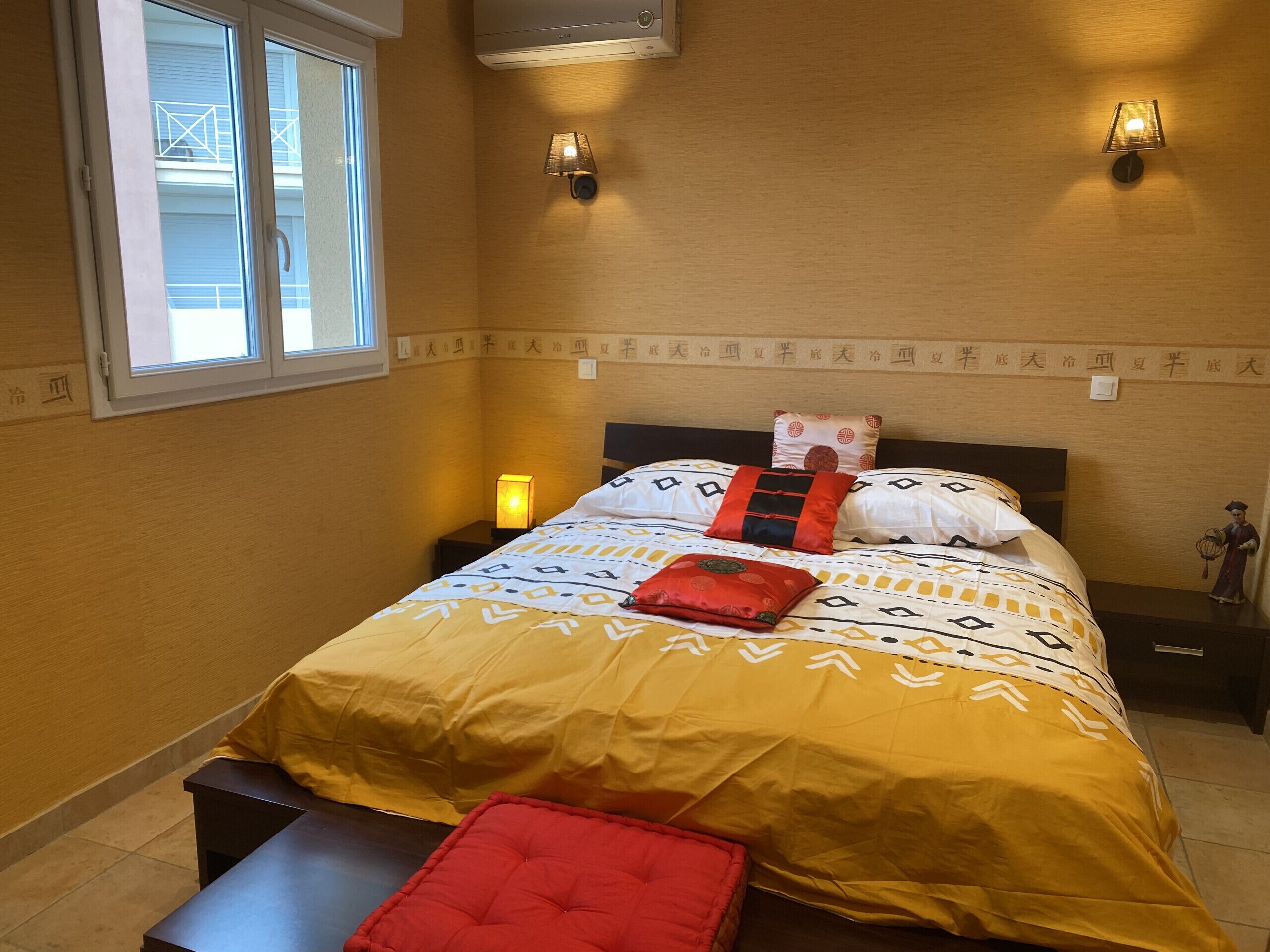 A hotel room with a bed, a nightstand, a bedside lamp, a wall-mounted air conditioner, and a window with a view of a balcony.