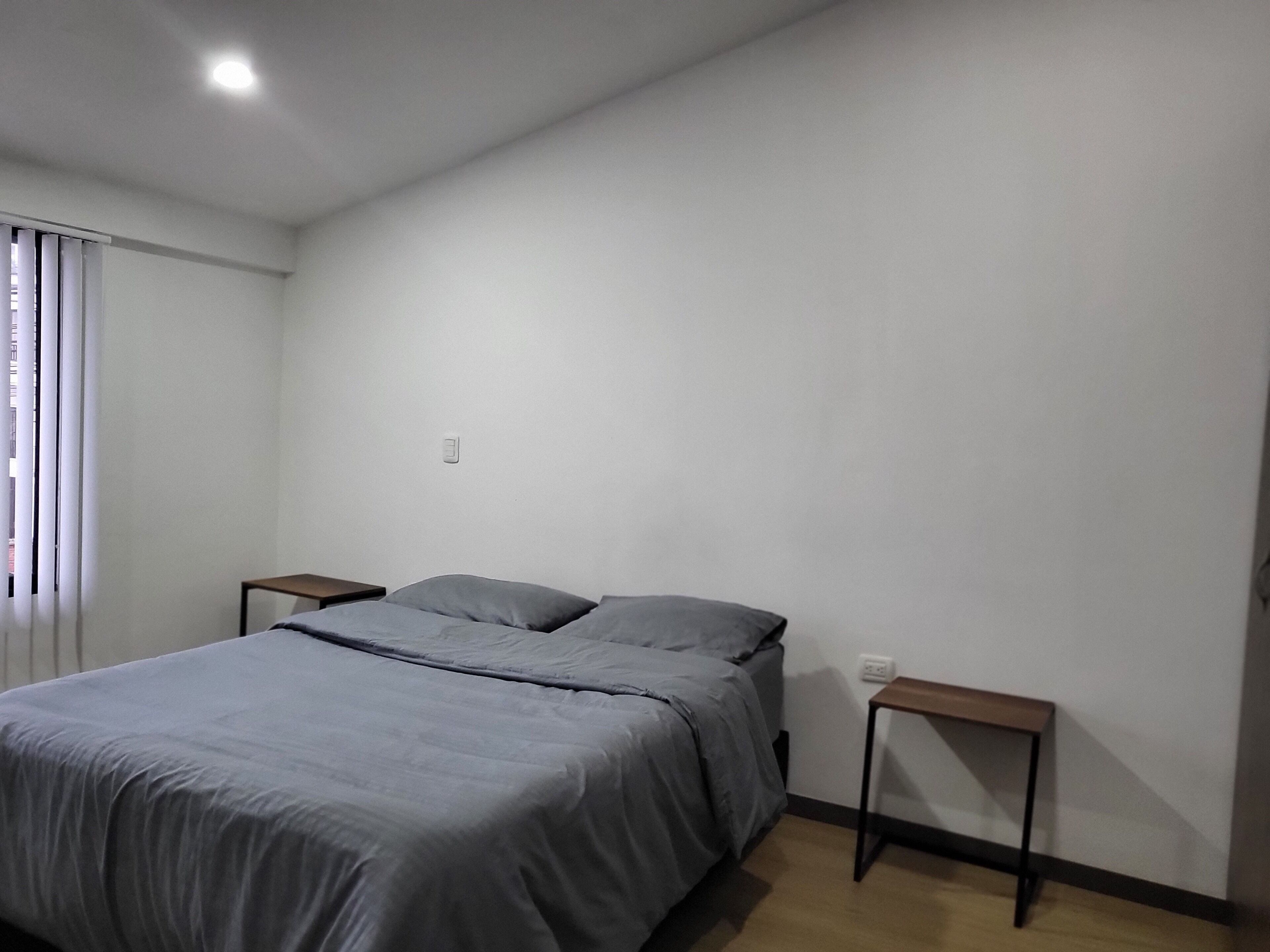 A bedroom with a bed, two nightstands, a window with blinds, and a light switch on the wall.