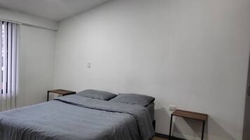A bedroom with a bed, two nightstands, a window with blinds, and a light switch on the wall.