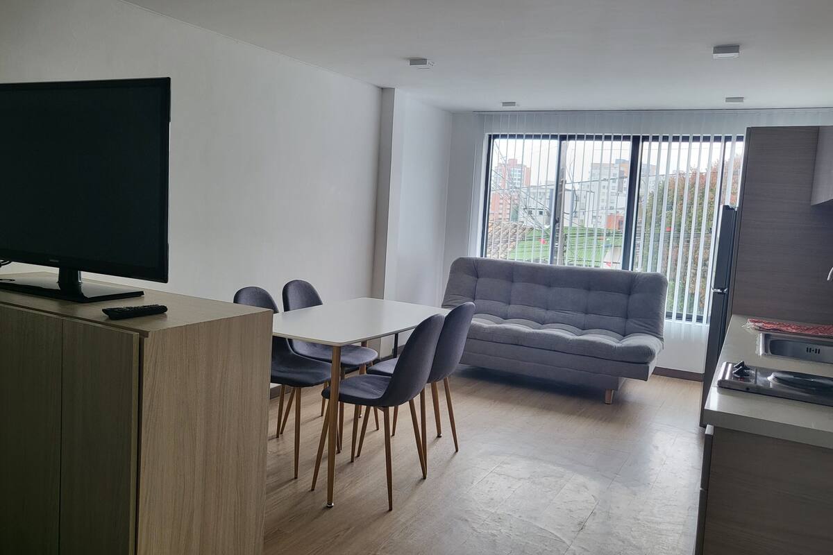 A modern living room with a flat-screen TV, a grey sofa, and a dining area with a table and chairs.