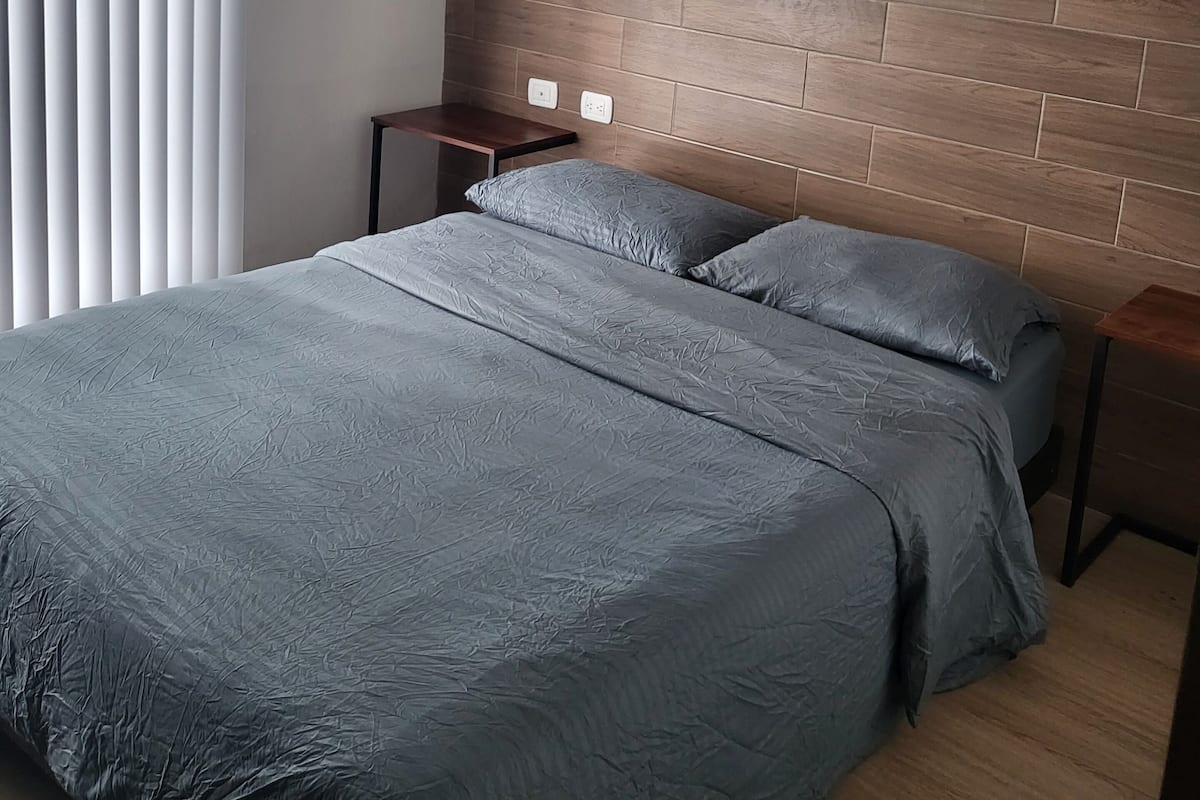 A bedroom with a wooden headboard, a bed with grey bedding, a nightstand, and a window with blinds.