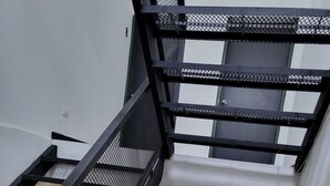 Un interior moderno con una escalera de metal negro que conduce a un segundo piso. Se observa un pasillo con piso de baldosas y una puerta al final.