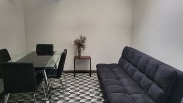 A room with a glass-top table, black chairs, a dark sofa, a small wooden side table with a vase, and a checkered floor.