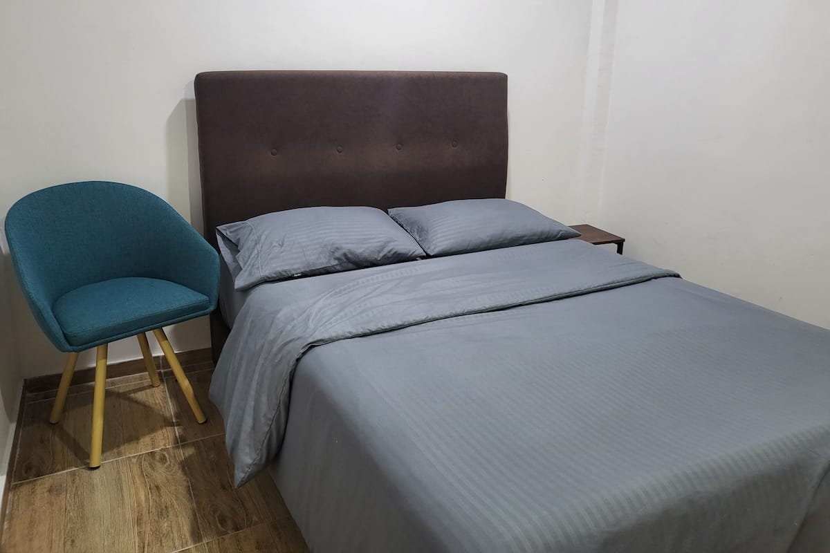 A bed with a dark headboard, two pillows, and a blue chair with wooden legs.