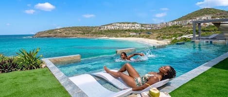 A pool with a view of the sea, a lounge area, and a hill with buildings in the background.