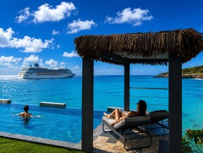 A pool area with lounge chairs and a thatched roof structure, overlooking a clear blue sea with a cruise ship in the distance. - Condostmaarten panoramic (Adults Only) (Sint Maarten)