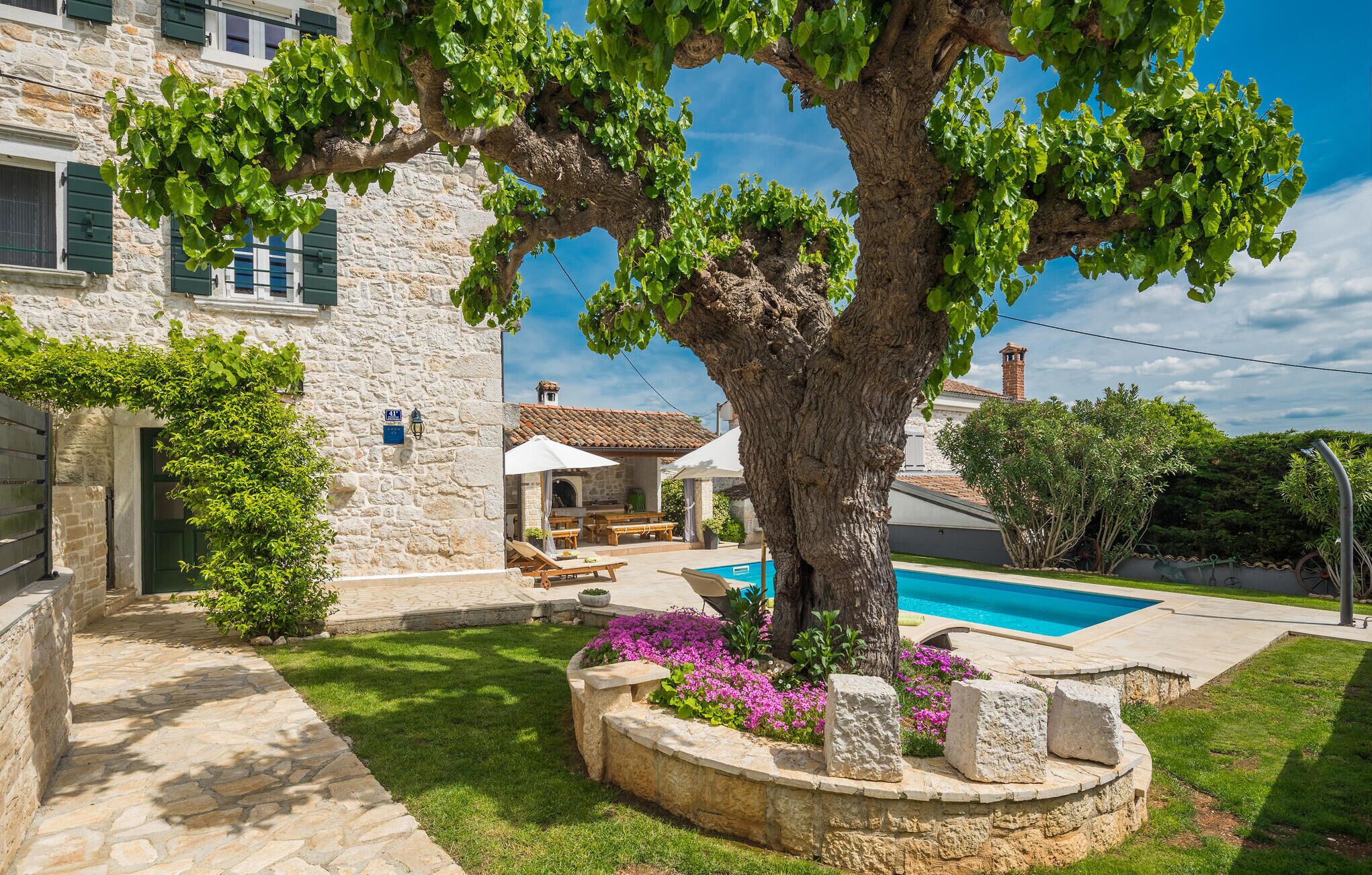 Una villa de piedra con piscina, mobiliario exterior y un gran árbol en el jardín.