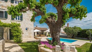 Una villa in pietra con piscina, area salotto esterna e un grande albero nel giardino.