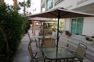 An outdoor patio area with tables and chairs, surrounded by potted plants and a white building in the background.