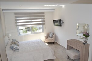 A modern bedroom with a large bed, a bedside table, a chair, a TV mounted on the wall, and a mirror above the vanity.