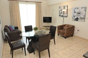 A modern living room with a glass dining table, dark leather chairs, a sofa, a patterned armchair, a flat-screen TV, and two large floral paintings on the wall.