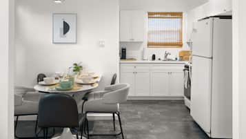 A modern kitchen with white cabinets, a dining table with chairs, a refrigerator, and a window with blinds.