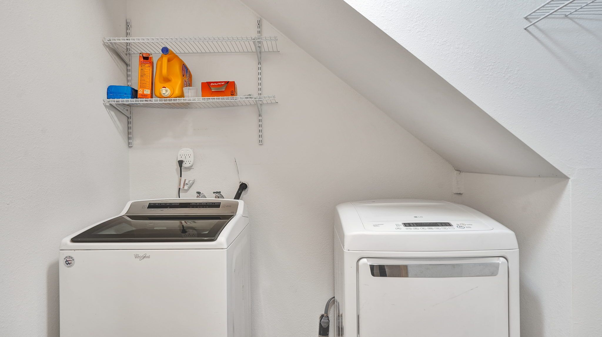 Un lavadero con lavadora y secadora Whirlpool, un estante con botellas de detergente y un enchufe.