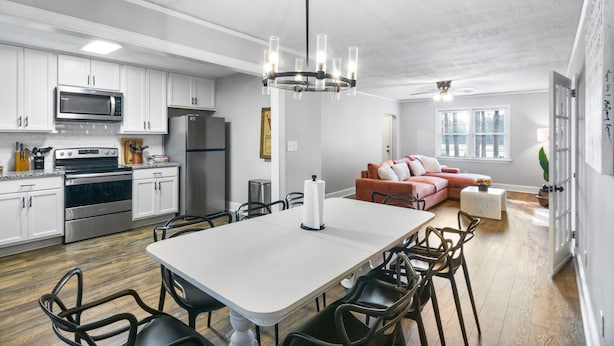 A modern kitchen with white cabinets, stainless steel appliances, and a dining area with a white table and chairs. - Forest Hills Cottage - 4.2 Miles from Masters (Augusta)