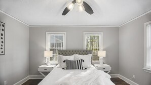 A bedroom with a large bed, two bedside tables with lamps, a ceiling fan, and two windows with blinds. - Forest Hills Cottage - 4.2 Miles from Masters (Augusta)