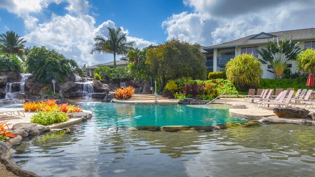A pool with a waterfall, surrounded by tropical plants and a building with a balcony.
